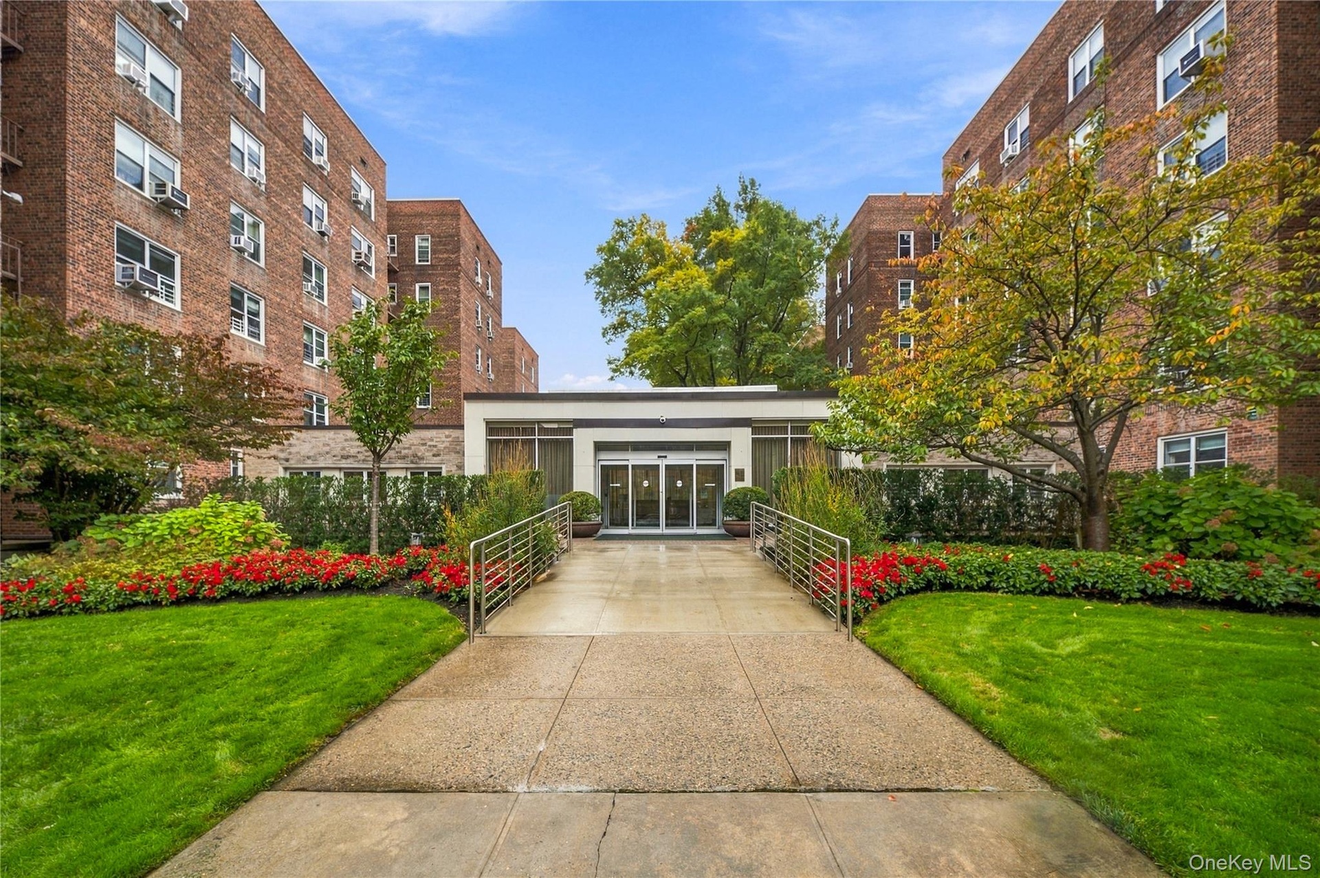 Brick masonry exterior featuring a central glass-paneled entryway, concrete walkway, and landscaped garden beds with mature trees