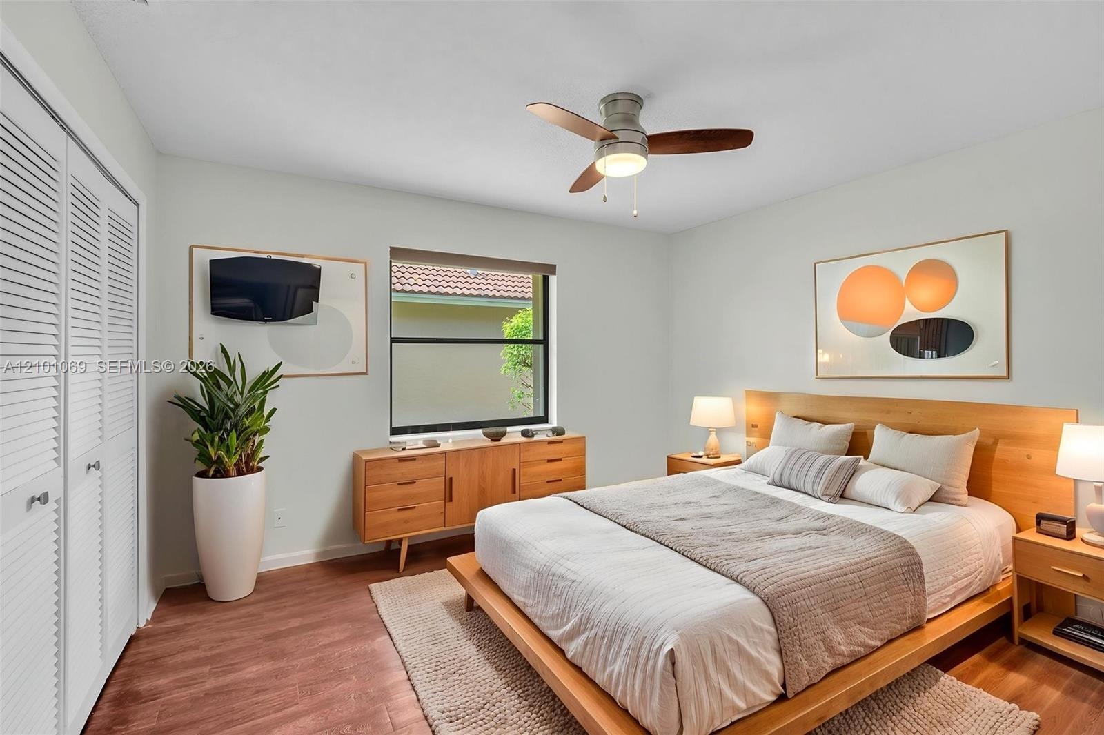Bedroom featuring wood-finish flooring, white louvered closet doors, a central ceiling fan with integrated lighting, and a window with a neutral rolle