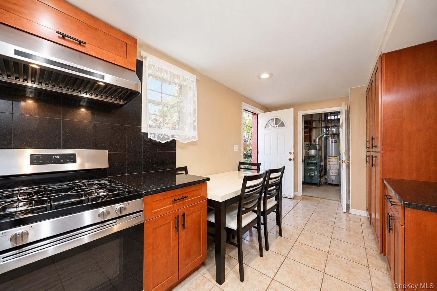 Kitchen featuring wood cabinetry, stone-finish countertops, tile backsplash, stainless steel gas range with a matching wall-mounted hood, and ceramic 