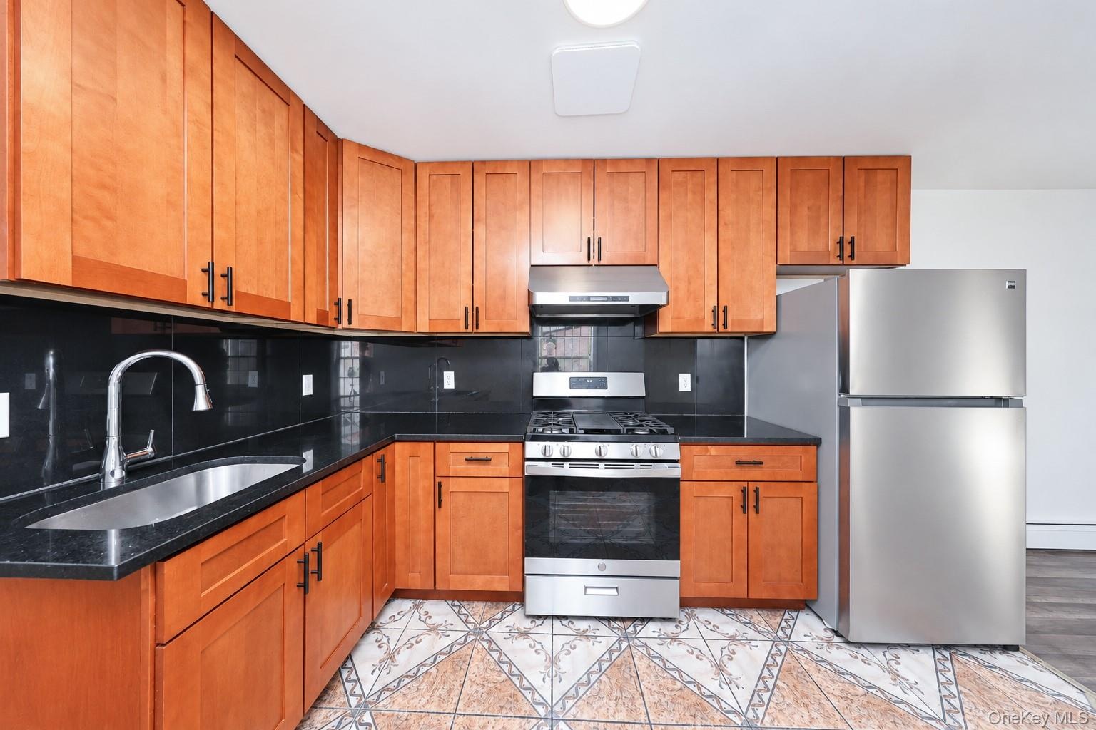 Kitchen featuring natural-finish wood cabinetry, black countertops, stainless steel appliances, and a geometric-pattern tile floor
