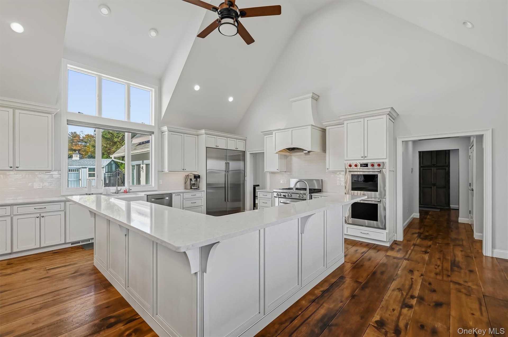 Kitchen featuring vaulted ceilings, wide-plank wood-finish flooring, white shaker-style cabinetry, a quartz surface peninsula, and stainless steel bui
