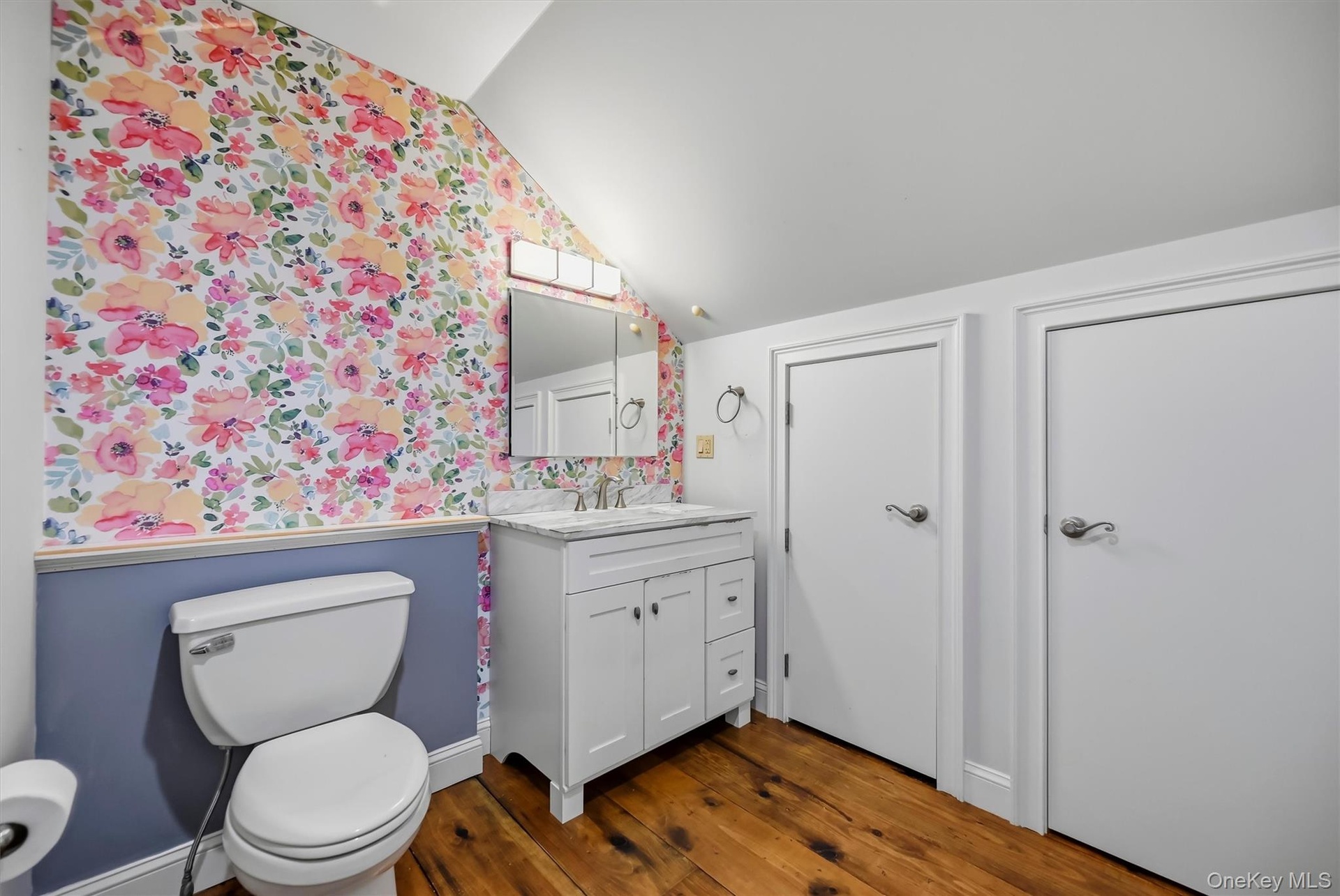 Bathroom featuring wood-finish flooring, floral accent wallpaper, wainscoting, a vanity with a light-toned stone countertop, and a built-in medicine c