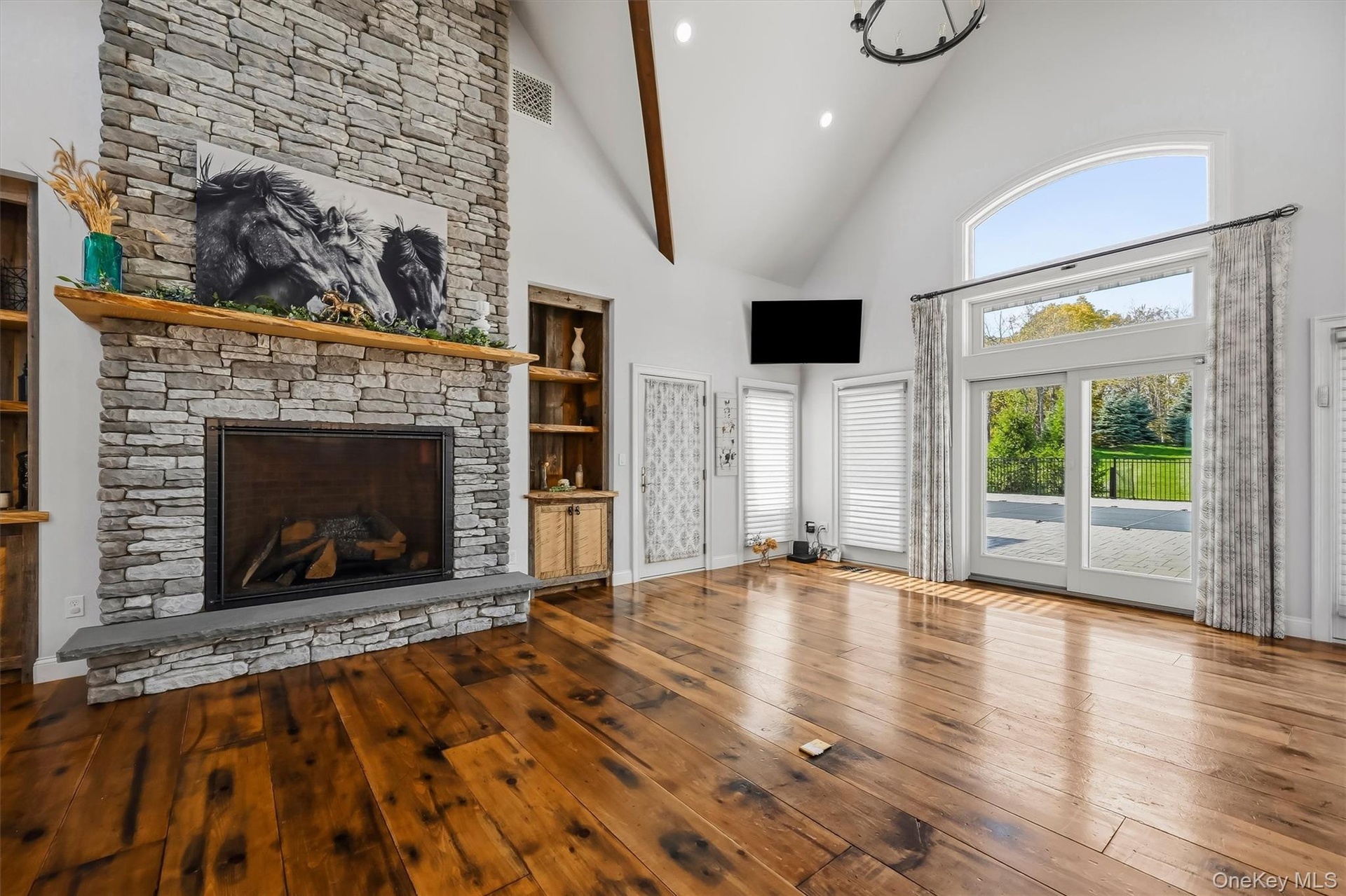 Vaulted ceilings with exposed beams, floor-to-ceiling stone  gas fireplace with wood mantel, wood-finish flooring, built-in shelving, and oversized gl