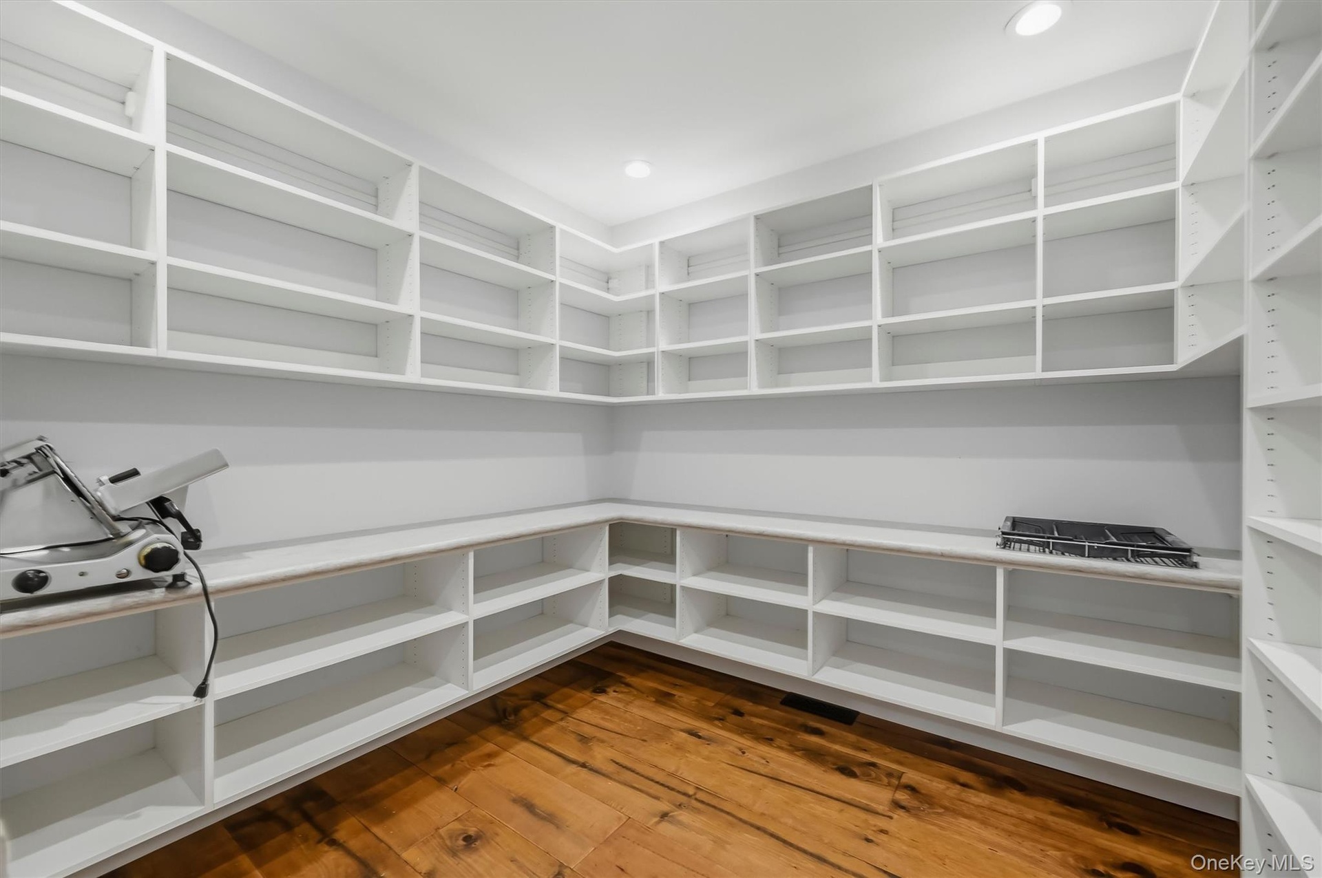 Walk-in pantry featuring floor-to-ceiling white built-in shelving, expansive solid surface countertops, and wide-plank wood-finish flooring