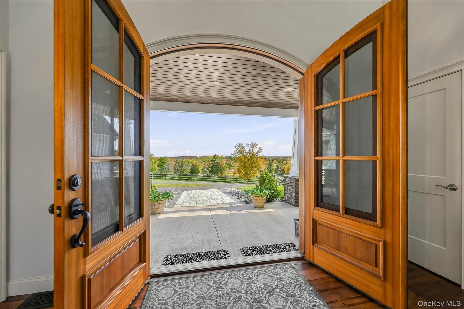 Arched double-entry natural wood doors with glass panels and matte black hardware, opening to a covered porch with a tongue-and-groove ceiling and pav