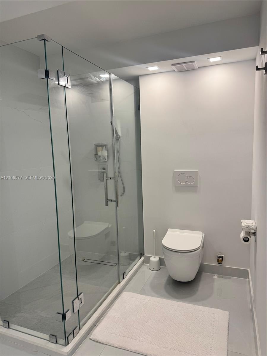 Frameless glass walk-in shower with stone-finish tiling, wall-mounted toilet, recessed ceiling lighting, and chrome fixtures