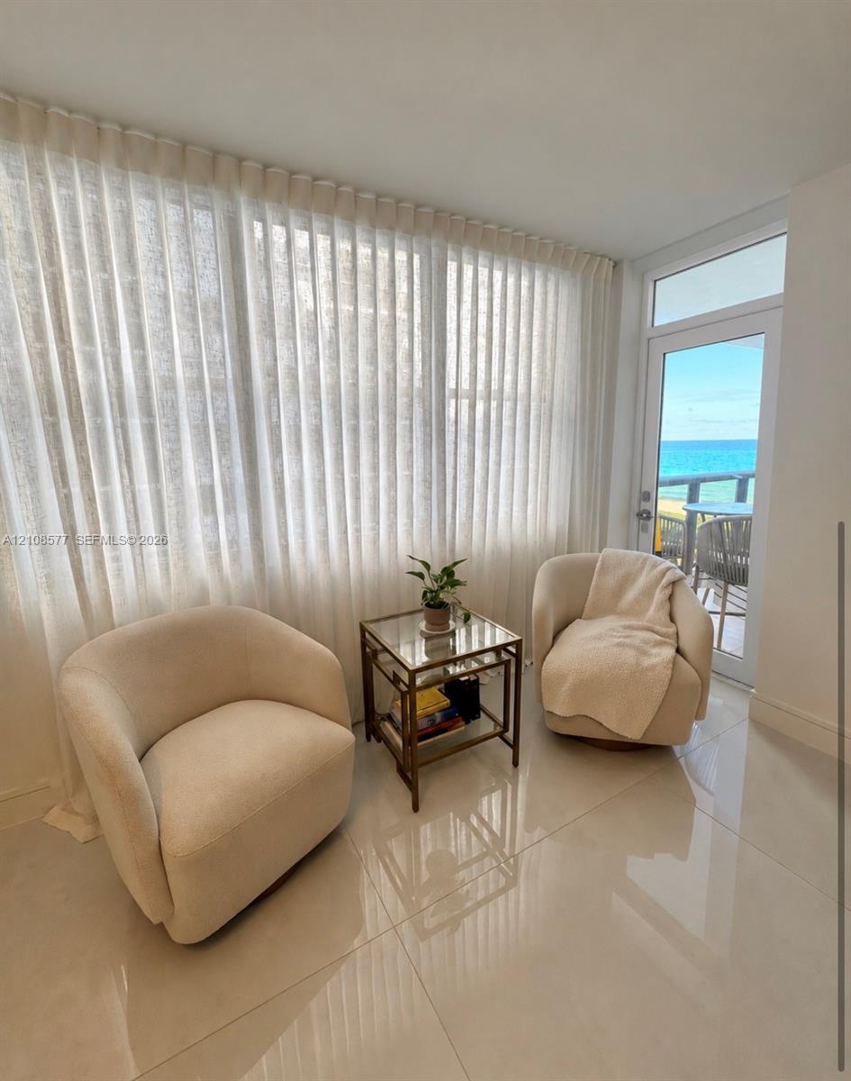 Floor-to-ceiling pleated window treatments, polished tile flooring, and a glass-paneled door leading to an outdoor terrace with ocean scenery