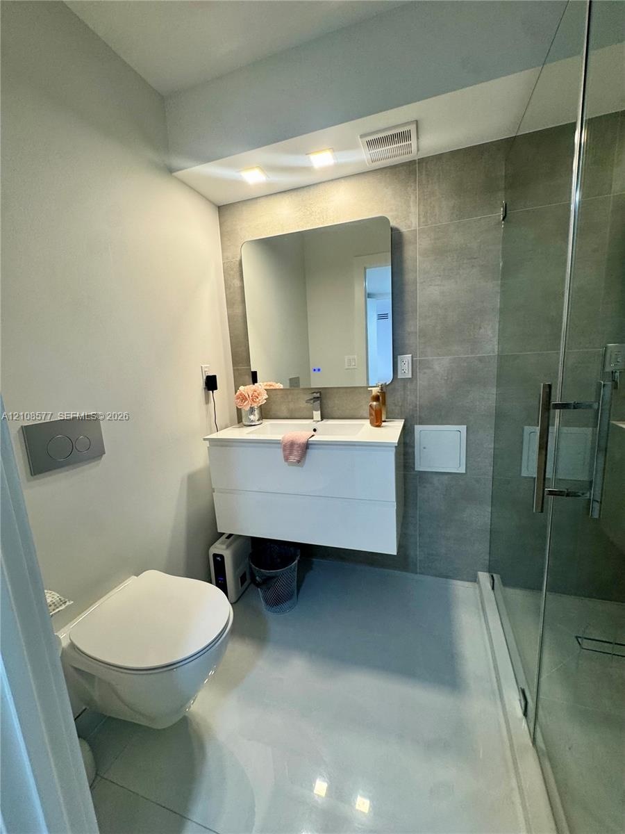 Floating vanity with a white countertop and integrated sink, wall-mounted toilet with a flush plate, and walk-in glass-enclosed shower with large-form