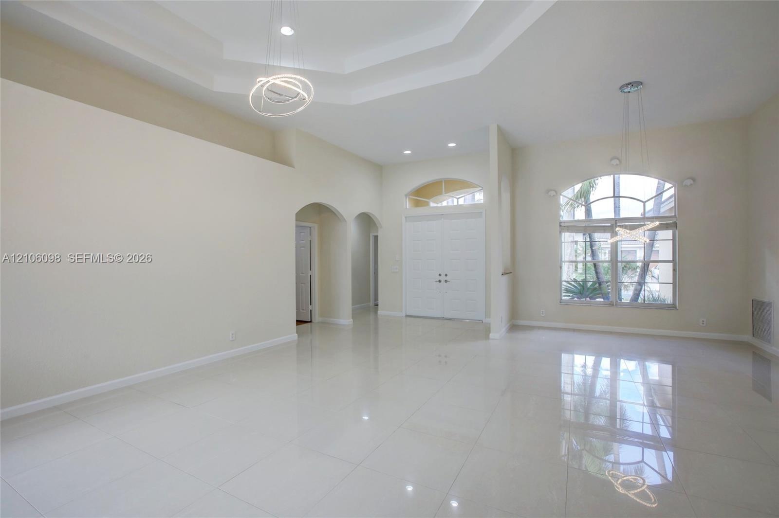 Open-concept interior featuring high tray ceilings, polished tile flooring, arched doorways, a double-entry door with transom window, and an expansive
