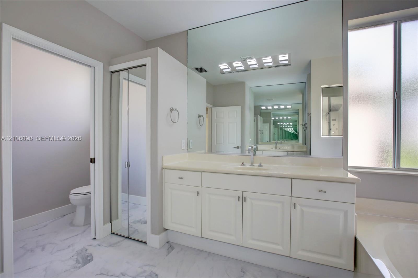 Bathroom featuring white cabinetry, solid-surface vanity countertop, full-width wall mirror, chrome-finish plumbing fixtures, and marble-patterned til