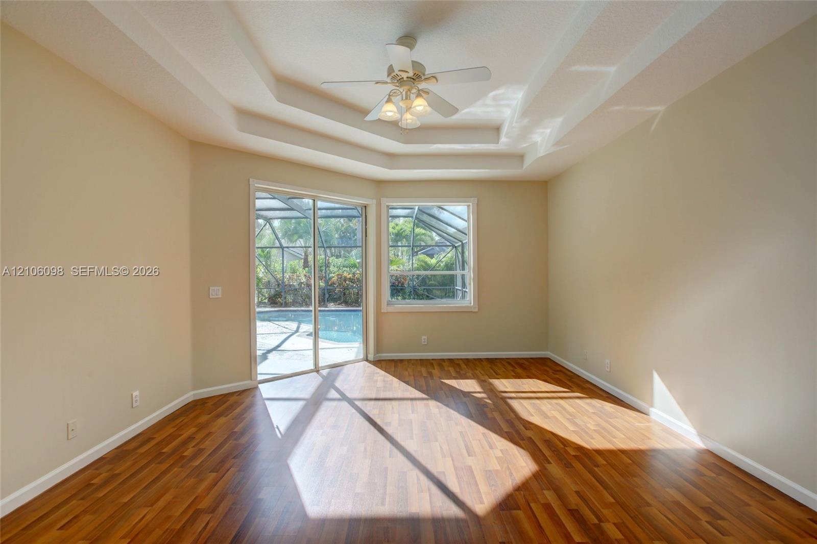 Room featuring tray ceilings, wood-finish flooring, a ceiling fan with integrated lighting, and sliding glass doors with access to a screened-in pool 