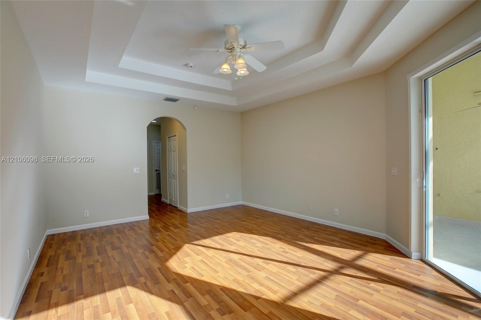 Room featuring a multi-tiered tray ceiling, wood-finish flooring, a central ceiling fan with multi-light fixture, an arched entryway, and a sliding gl