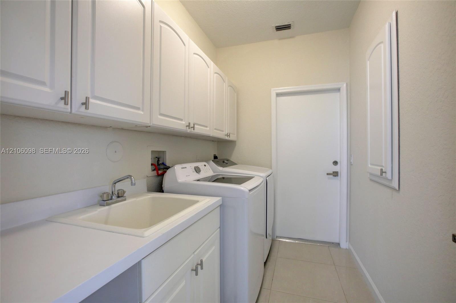 Utility area featuring white shaker-style wall cabinetry, a composite utility sink with a chrome faucet, solid-surface countertops, and tile flooring