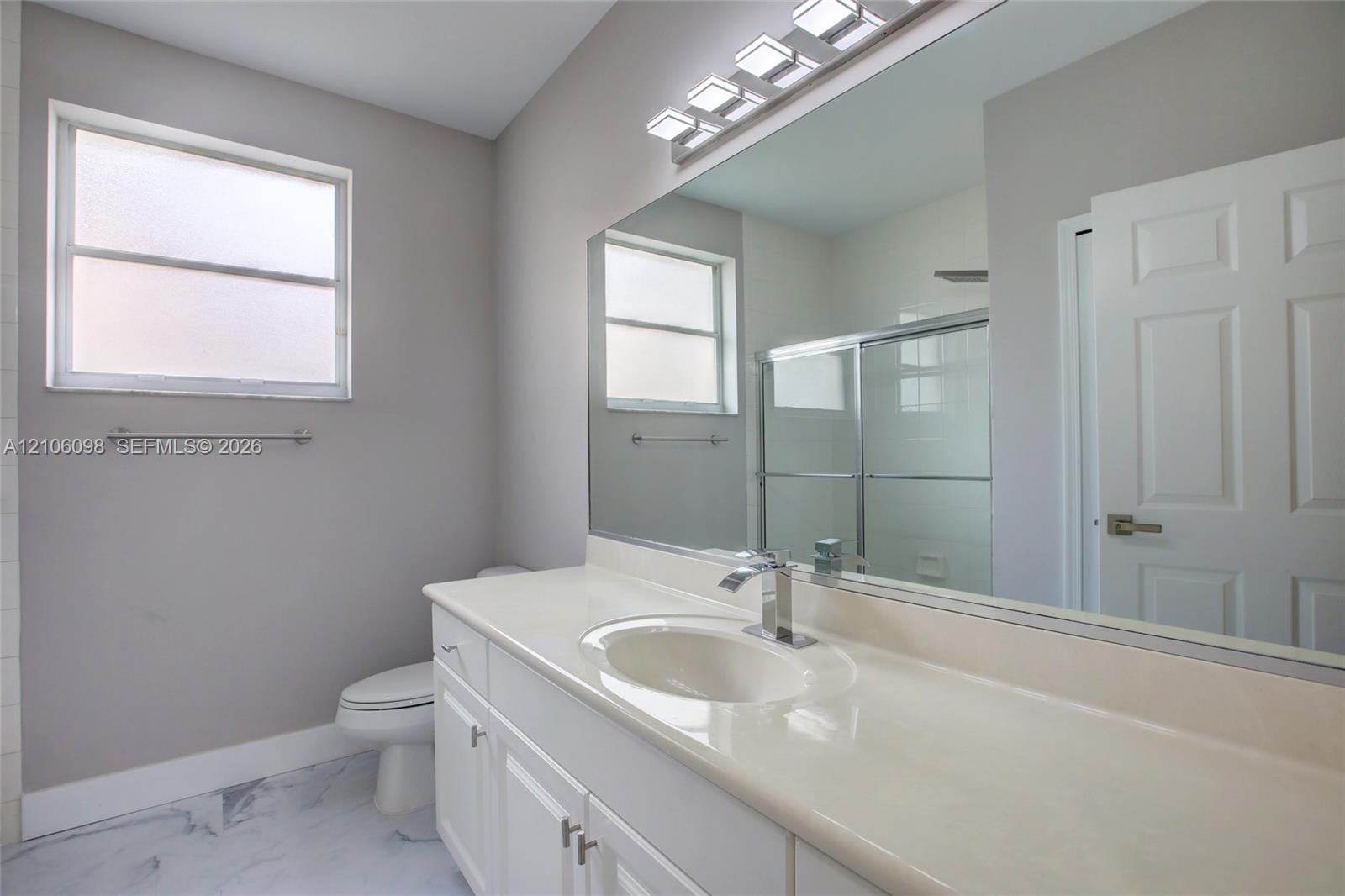 Bathroom featuring a solid-surface countertop with an integrated basin, white shaker-style vanity cabinetry, a full-width wall mirror, a multi-bulb li