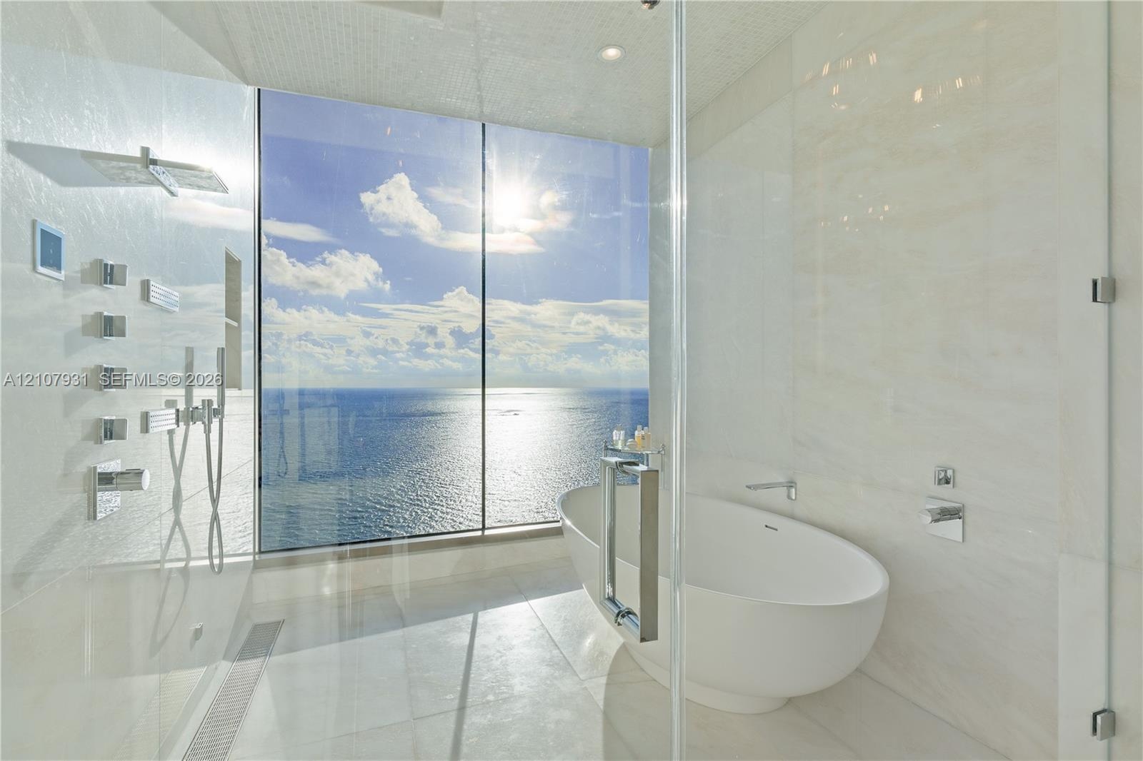 Floor-to-ceiling glass paneling, freestanding soaking tub, walk-in shower with multi-head wall-mounted shower system, and light-toned tile flooring