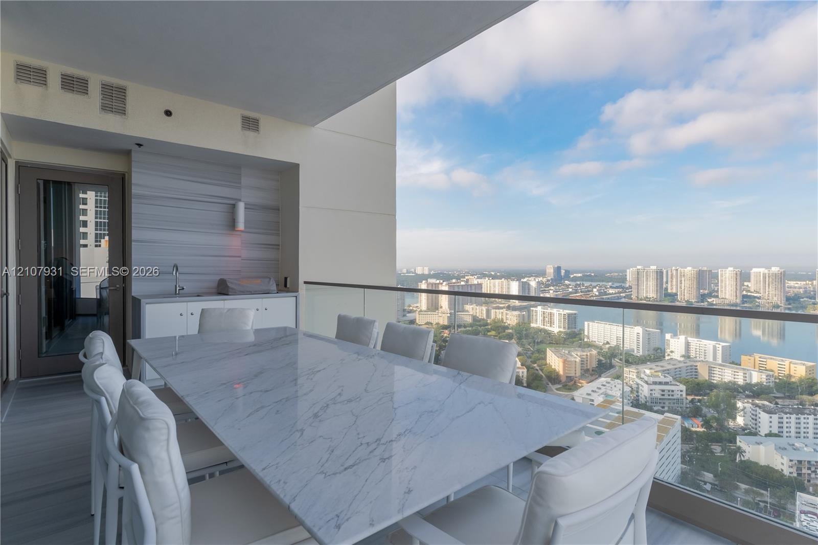 Covered balcony featuring an integrated outdoor kitchen with white cabinetry, horizontal tile wall accent, glass railing, and expansive water and city