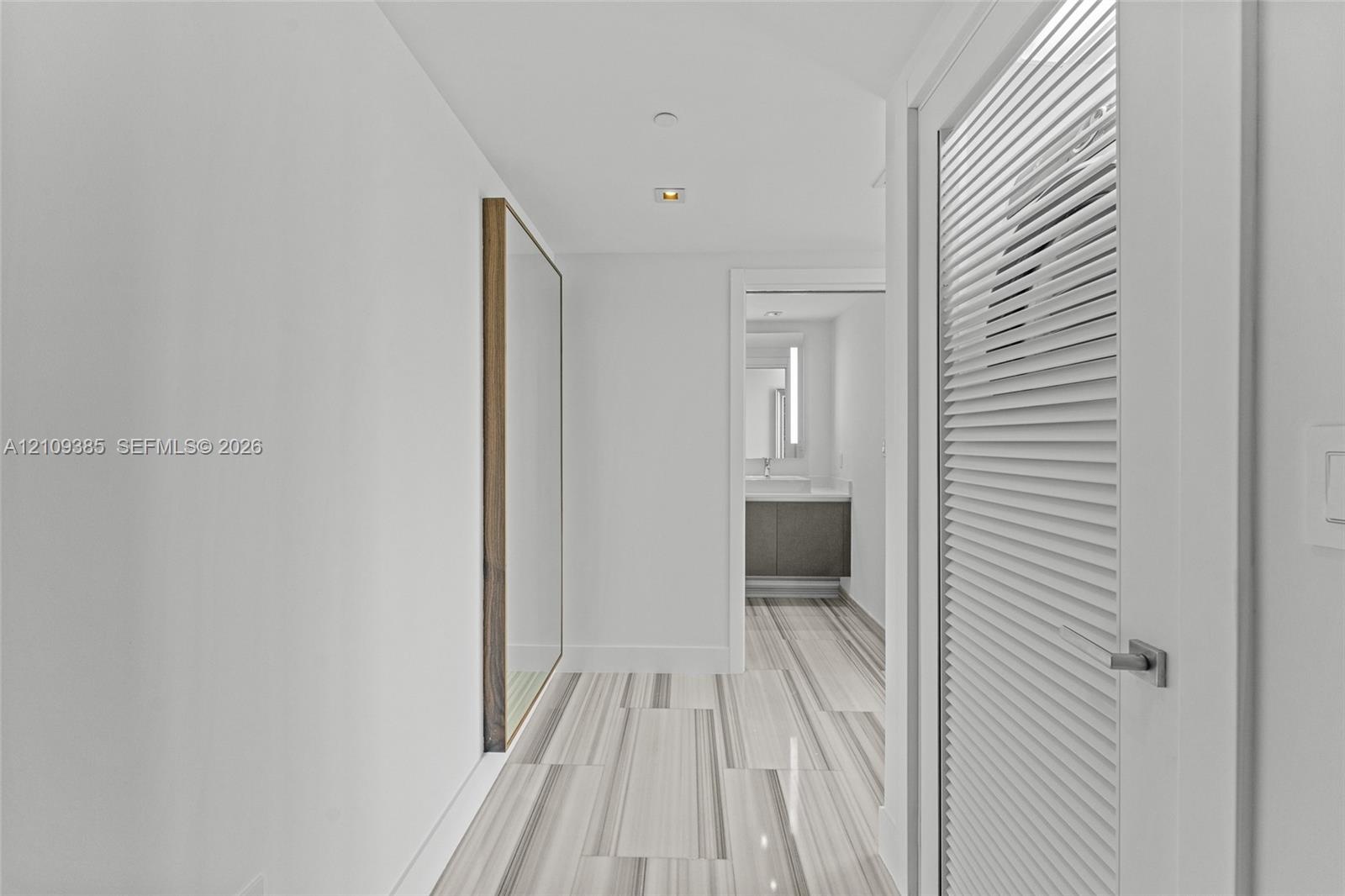 Hallway featuring linear-patterned tile flooring, a louvered white door with satin hardware, recessed ceiling lighting, and a flush-mounted wall mirro
