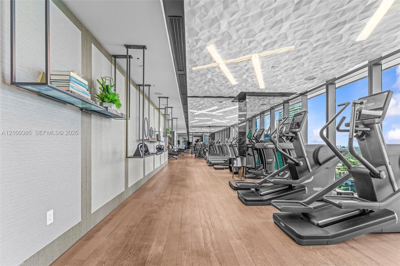 Fitness center featuring wood-finish flooring, sculptural geometric ceiling panels, linear recessed lighting, floor-to-ceiling windows, and wall-mount