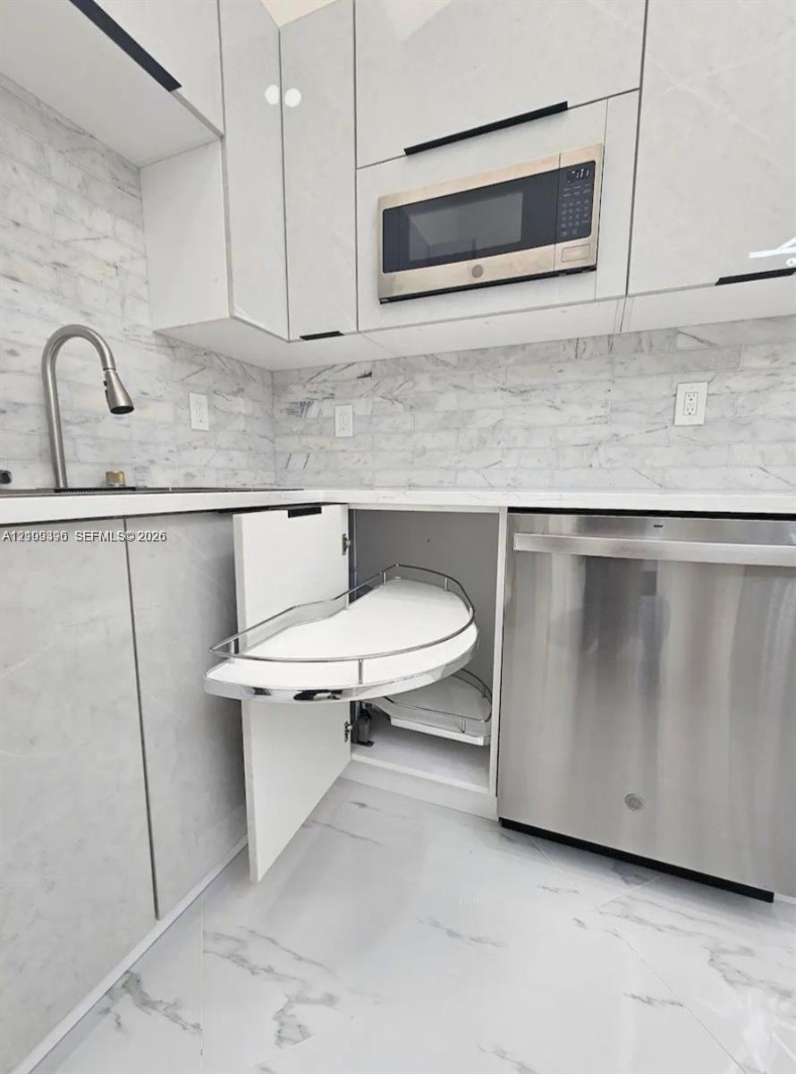 Modern kitchen featuring white flat-panel cabinetry, pull-out corner shelving, stainless steel appliances, stone-patterned backsplash, and matching st