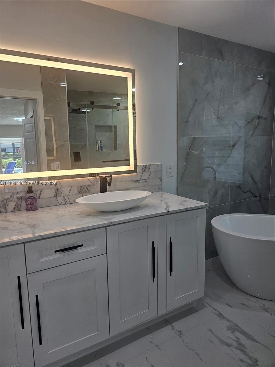 Modern white cabinetry with black hardware, stone-surface countertop, elliptical vessel sink, integrated LED mirror, and soaking tub with large-format