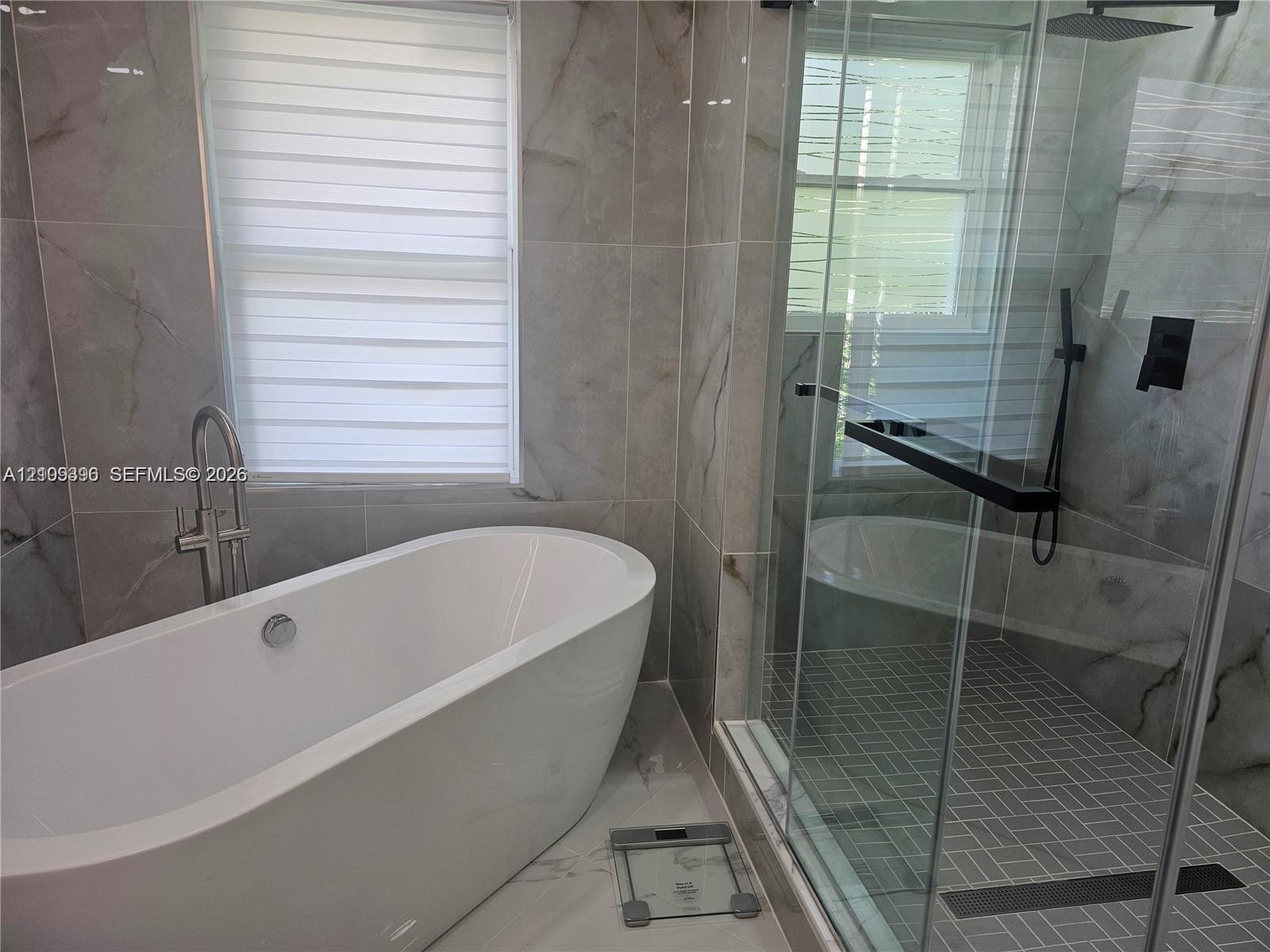 Freestanding oval soaking tub with floor-mounted faucet, walk-in glass-enclosed shower featuring a rainfall showerhead, and large-format wall tiling w
