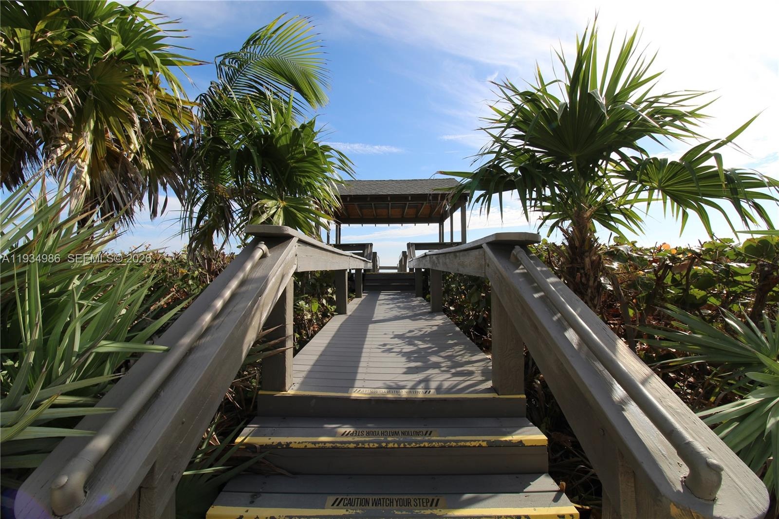 Elevated wooden boardwalk featuring handrails, flanked by mature palm trees and coastal vegetation, leading to a covered wooden pavilion