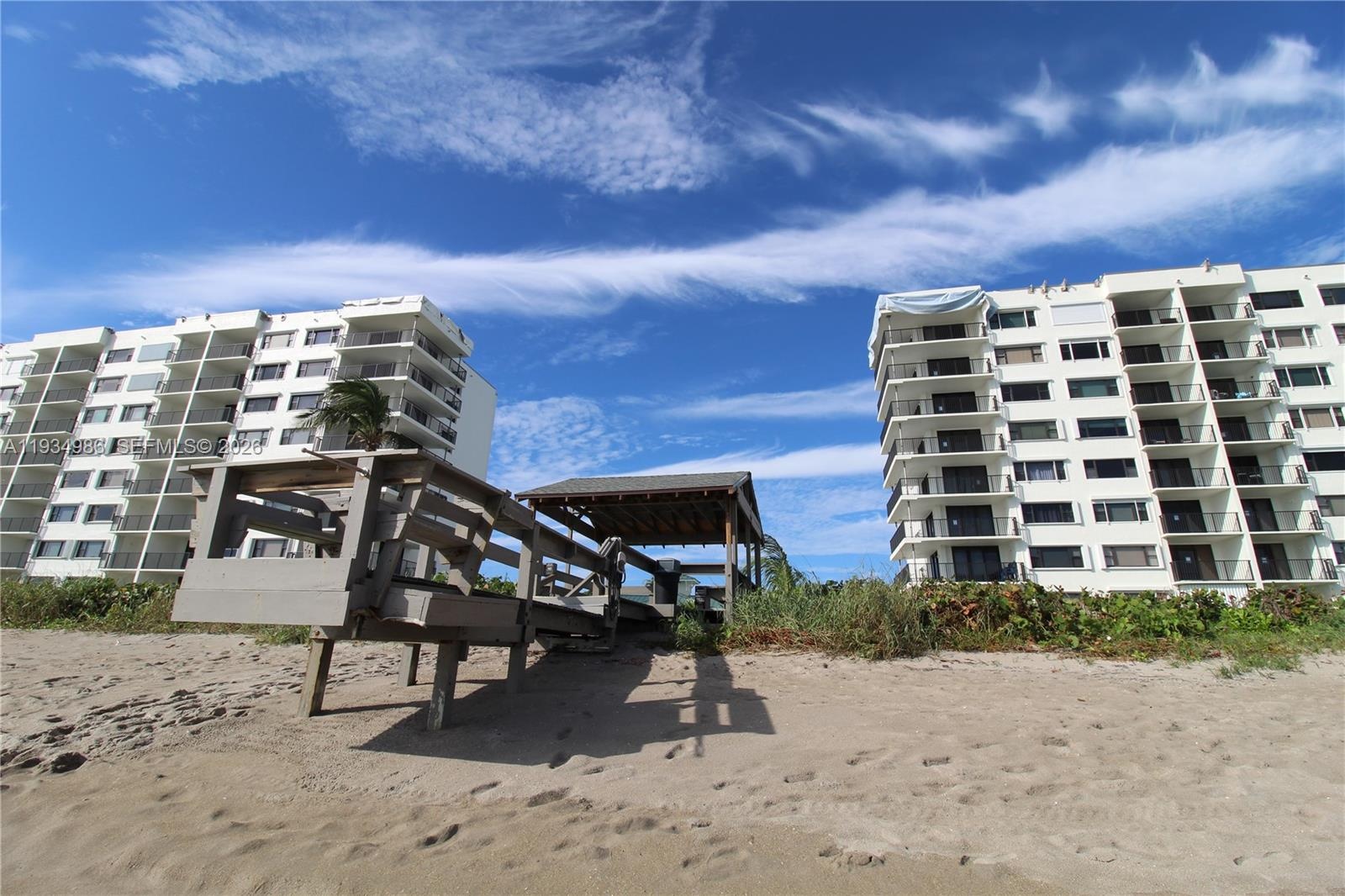 Exterior view of multi-story buildings featuring private balconies and a wooden boardwalk with a covered gazebo structure situated on sandy beach terr