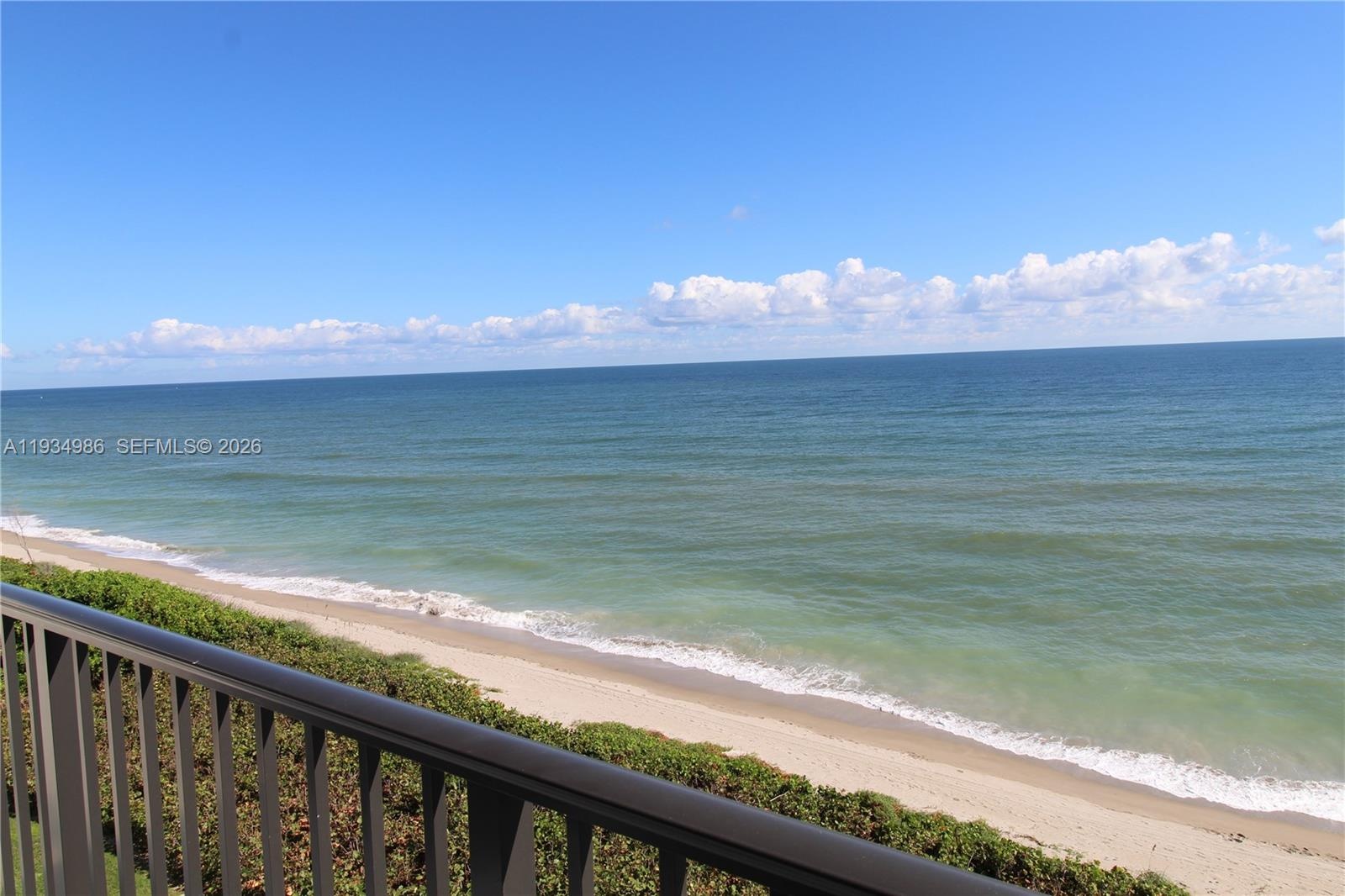 Private balcony featuring metal railings overlooking a sandy shoreline, ocean water, and horizon