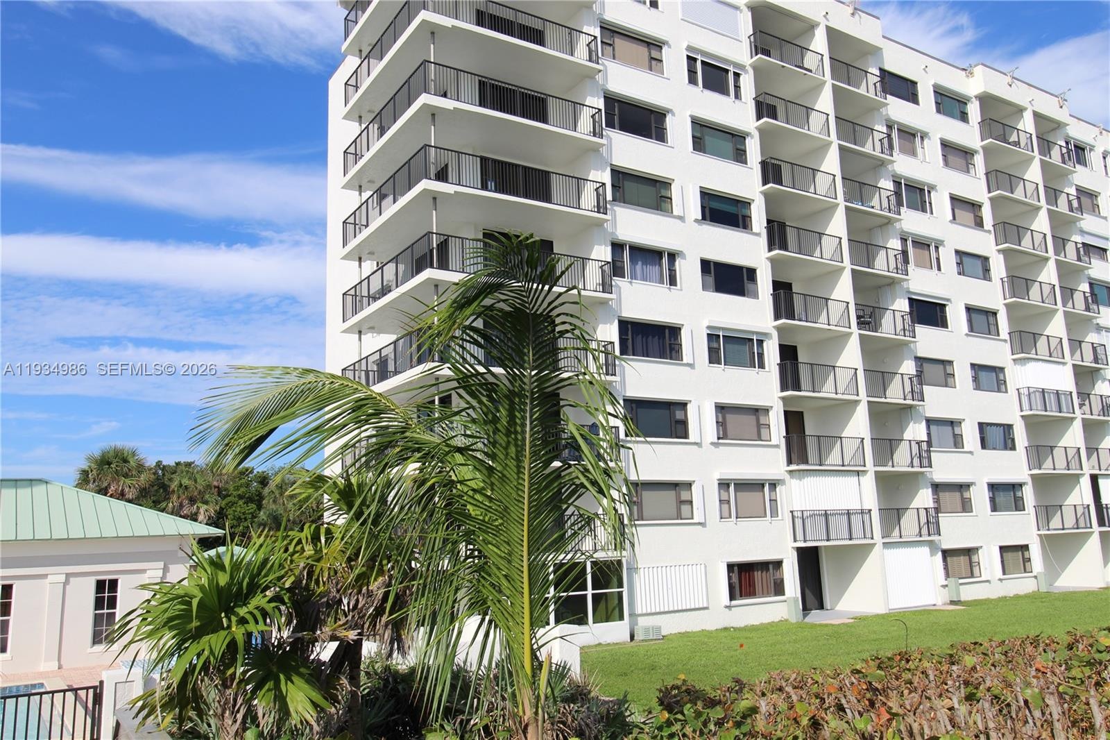 High-rise building exterior featuring tiered concrete balconies with metal railings, horizontal windows, and landscaped grounds