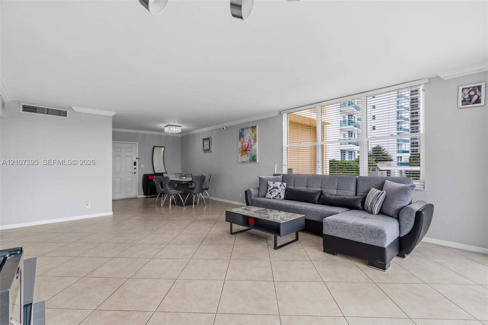 Open-concept living space featuring neutral ceramic tile flooring, crown molding, and floor-to-ceiling windows with horizontal blinds