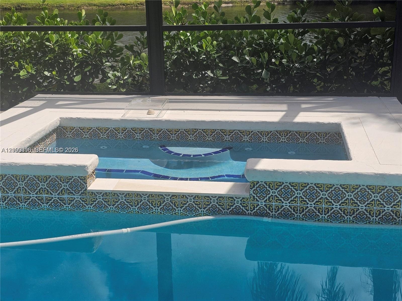 Raised spa featuring patterned decorative tile work and stone-style coping, enclosed within a screened lanai overlooking a water feature