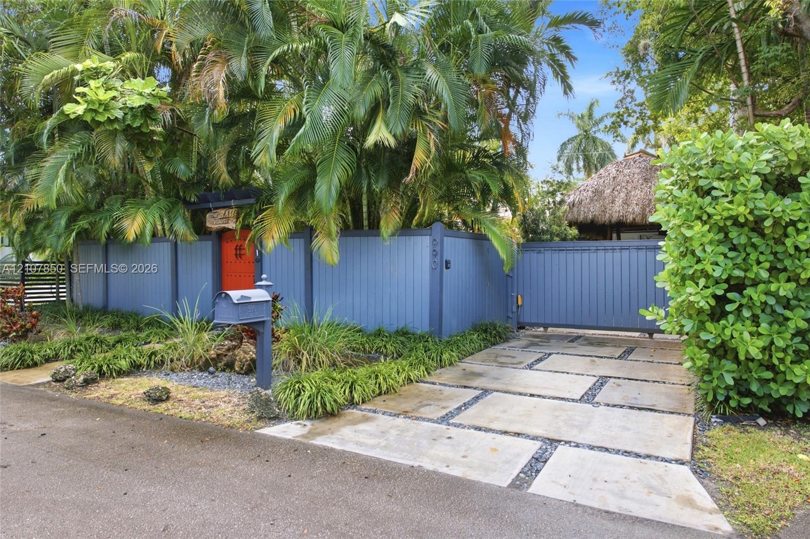 Private gated entry featuring a custom blue vertical-board fence, concrete paver driveway with crushed stone detailing, and mature tropical landscapin