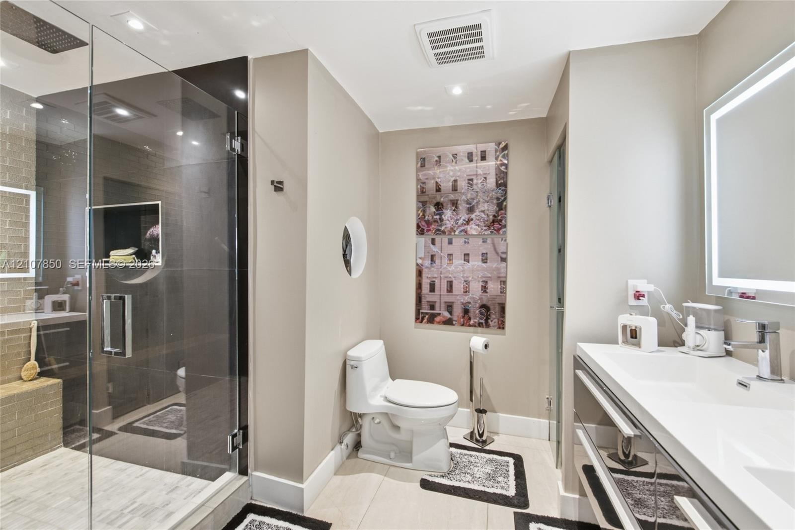 Glass-enclosed walk-in shower with stone tile flooring, modern dual-vanity with integrated sink, illuminated mirror, and custom tile wall accents