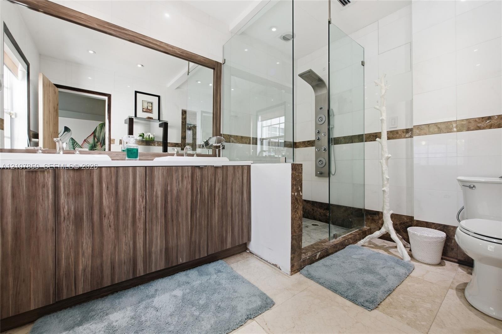 Dual-sink vanity with wood-finish cabinetry, full-width mirror, walk-in shower with frameless glass enclosure, and stone-tile flooring
