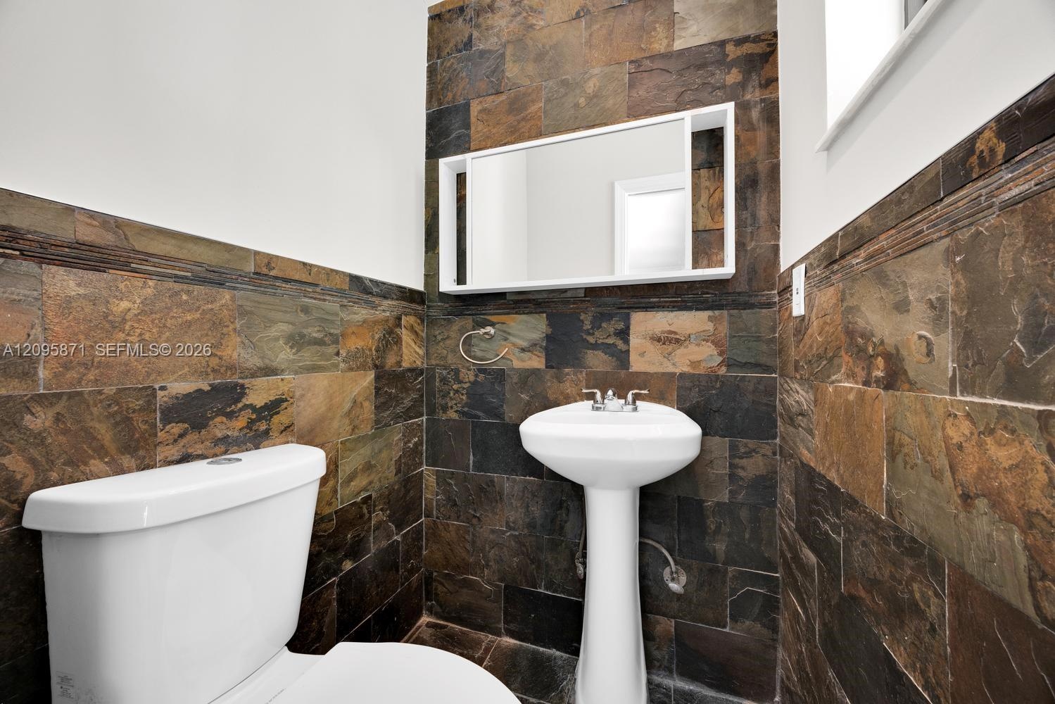 Powder room featuring natural stone tile wainscoting, a white pedestal sink with a chrome faucet, and a wall-mounted rectangular medicine cabinet
