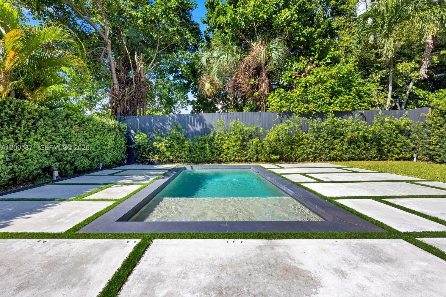 Rectangular swimming pool featuring a tanning ledge, bordered by concrete pavers with grass inlay, and enclosed by a solid privacy fence and manicured