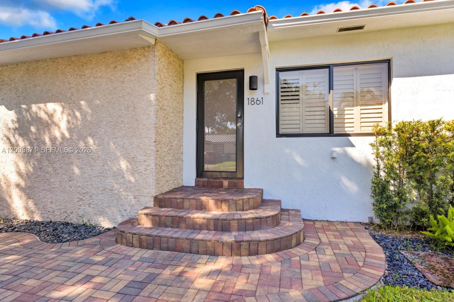 Exterior entrance featuring a terracotta tile roof, stucco facade, multi-level brick steps, multi-tone paver walkway, and a window with exterior plant