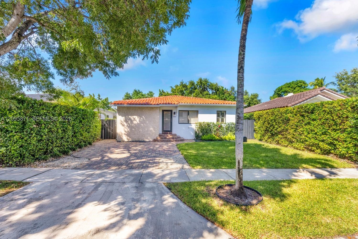 Single-story residence featuring a tile roof, light-toned stucco exterior, paver driveway, and dense perimeter hedging