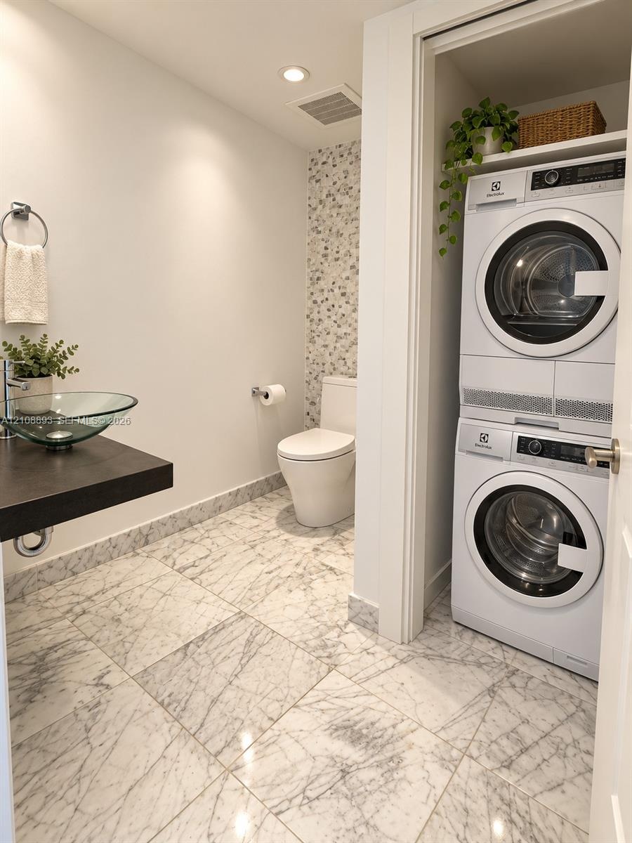 Full-bathroom featuring large-format stone-patterned floor tile, glass vessel sink with dark floating vanity, integrated laundry closet, and decorativ