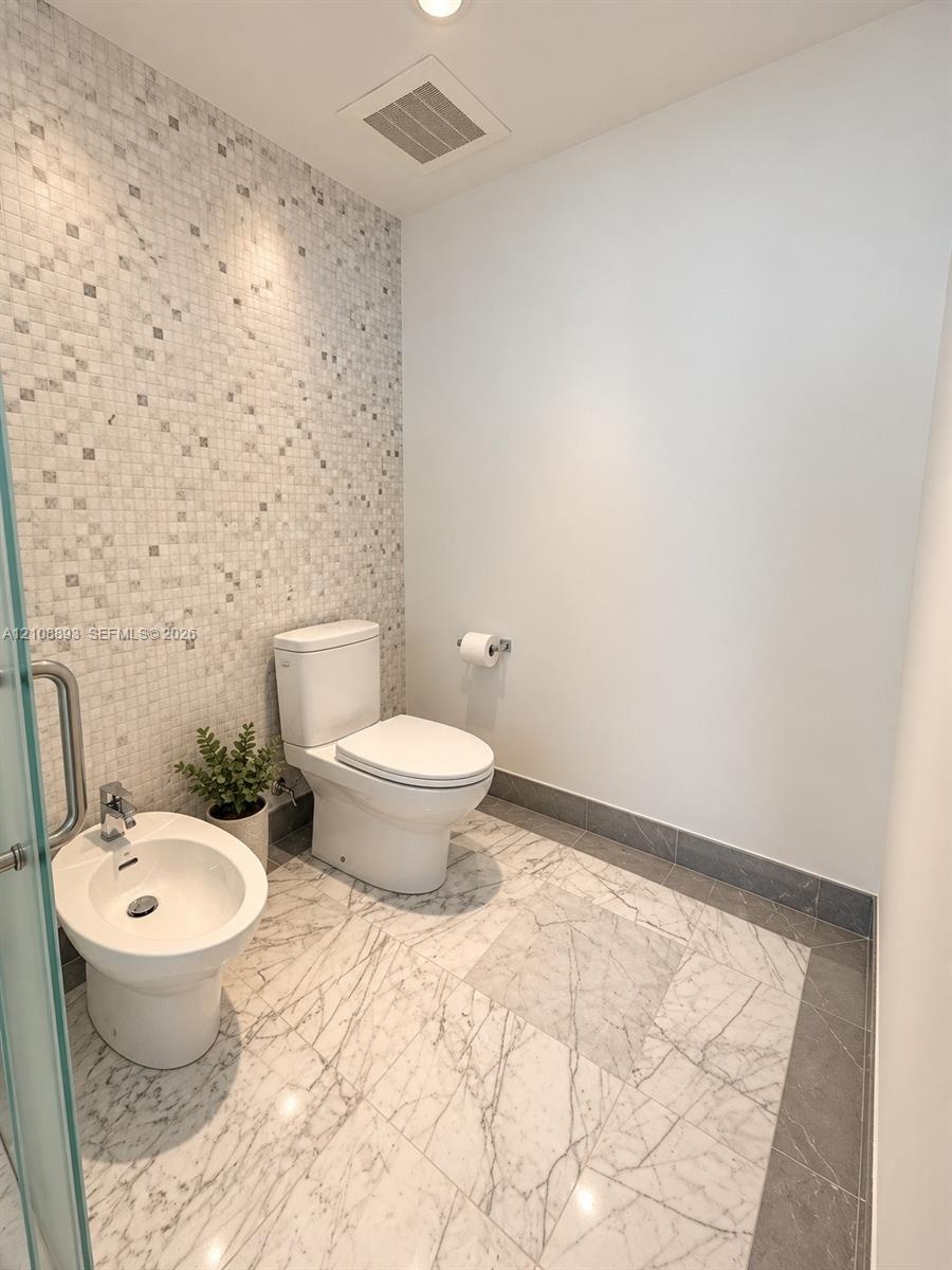 Wall-mounted bidet with chrome faucet, floor-mounted elongated toilet, mosaic tile accent wall, and stone tile flooring with border inlay