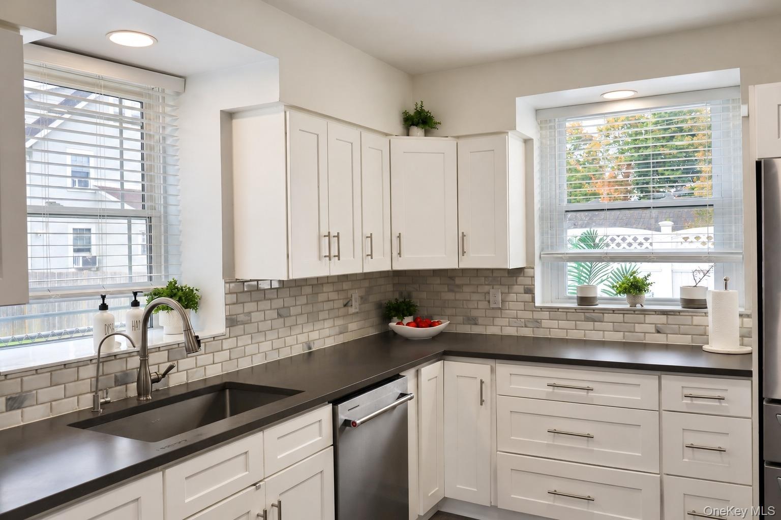 Kitchen featuring shaker-style cabinetry, dark solid-surface countertops, subway tile backsplash, stainless steel appliances, and dual windows