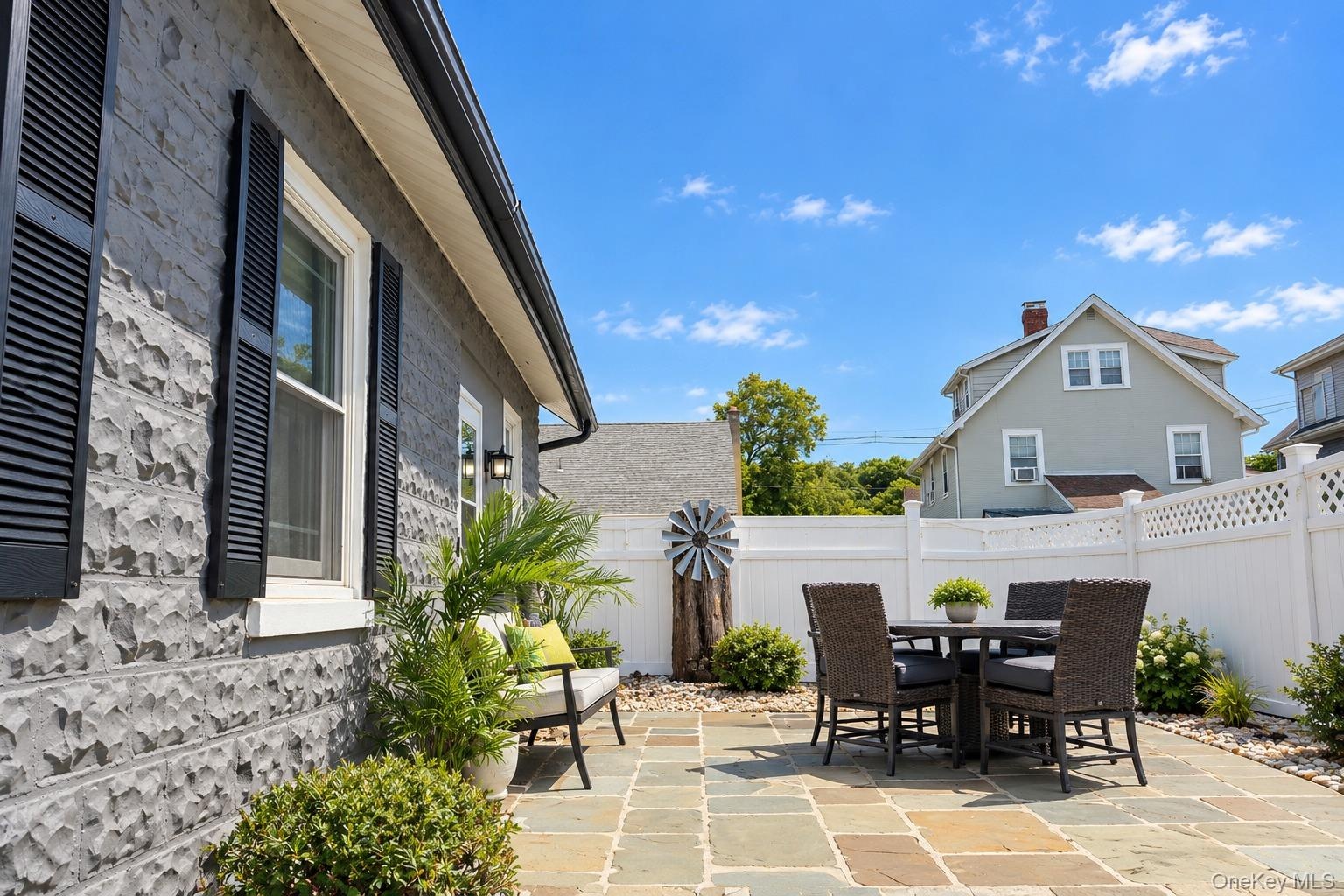 Exterior featuring grey rock-faced masonry siding, dark window shutters, natural stone paver patio, white vinyl privacy fencing, and metal decorative 