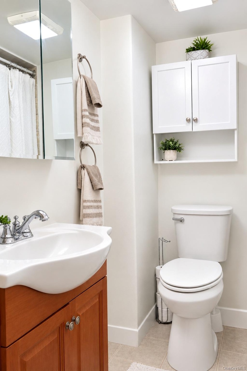 Wall-mounted white cabinetry with open shelving, wood-finish vanity, polished chrome faucet, rectangular wall-mounted mirror, and standard-height toil