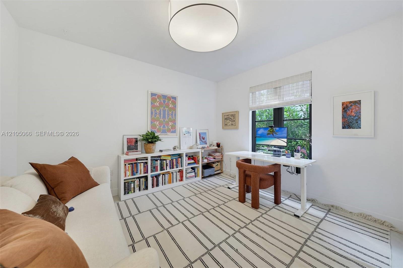 Neutral-toned room featuring a drum-style flush mount ceiling light, a dark-framed multi-pane window, and a woven roller shade