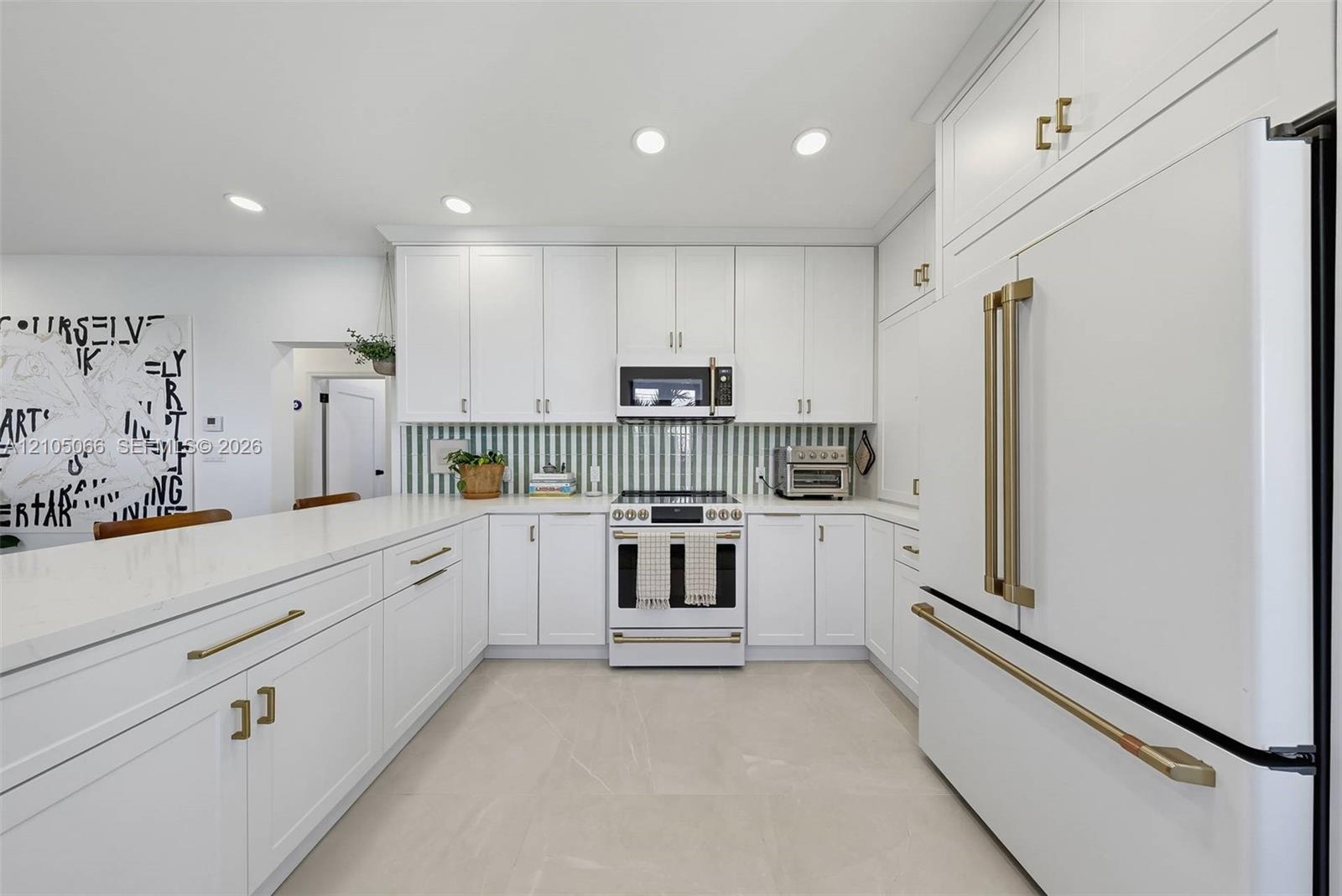 Kitchen featuring white shaker-style cabinetry, quartz countertops, a green and white vertical tile backsplash, gold-toned hardware, and white-finish 