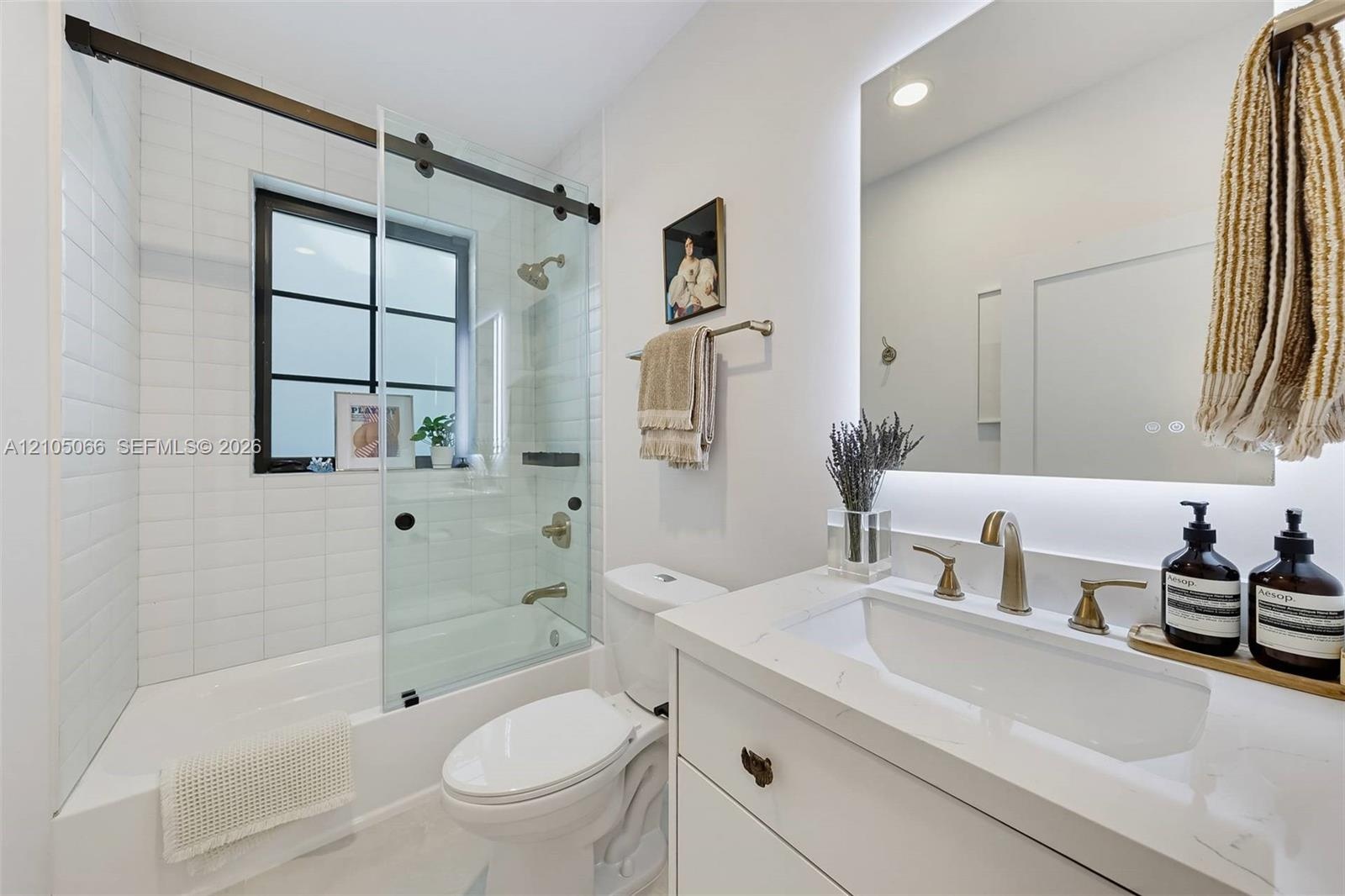 Bathroom featuring a vanity with a white countertop and brass-finish widespread faucet, a backlit mirror, a tub-shower combination with a sliding glas