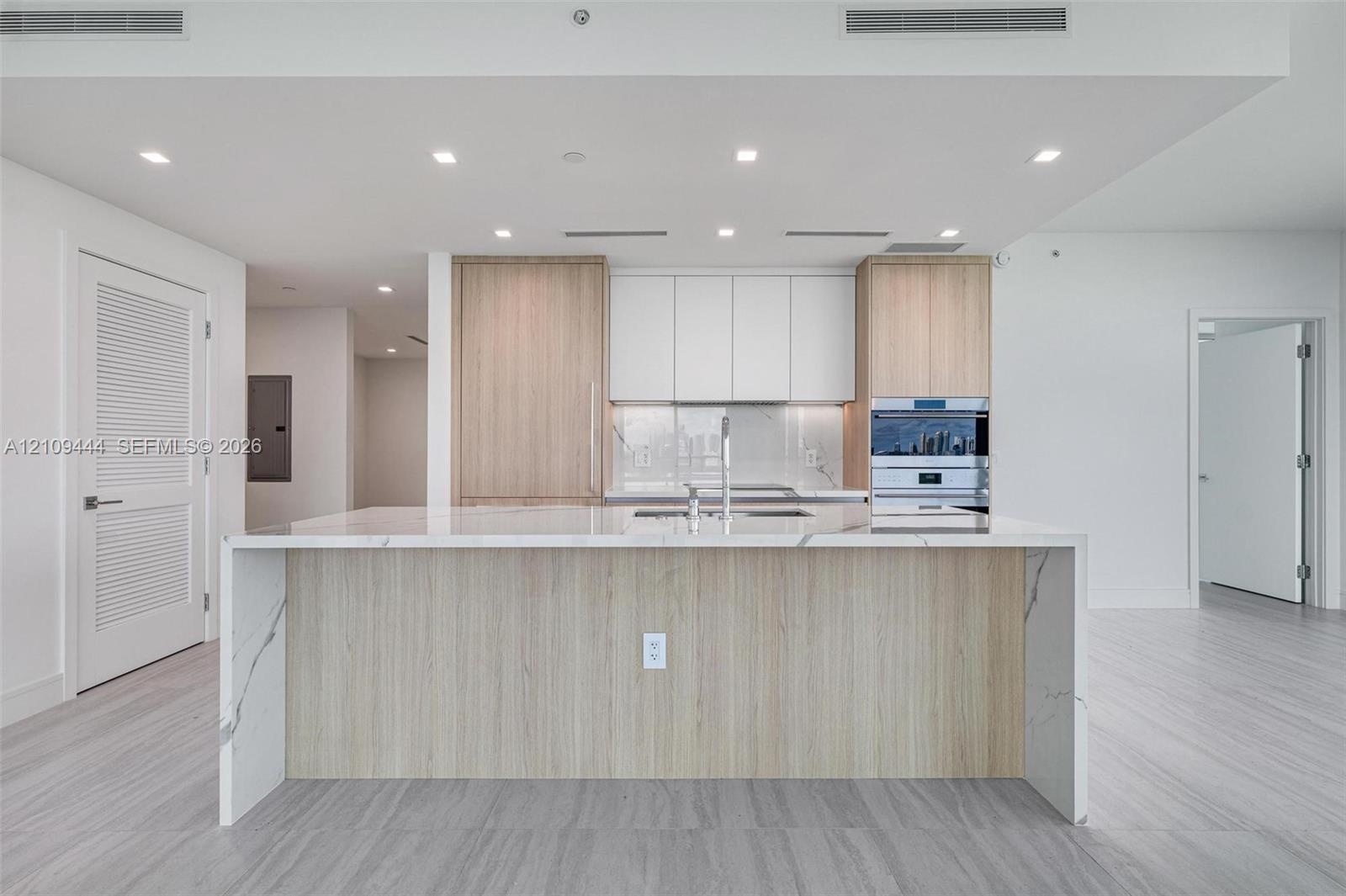 Kitchen featuring a center island with a stone-finish waterfall countertop, custom wood-finish cabinetry, white matte upper cabinets, integrated stain