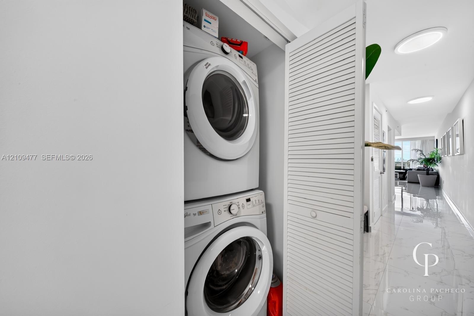 Laundry closet featuring stacked front-load appliances and louvered folding doors, accented by polished stone-finish flooring and recessed lighting