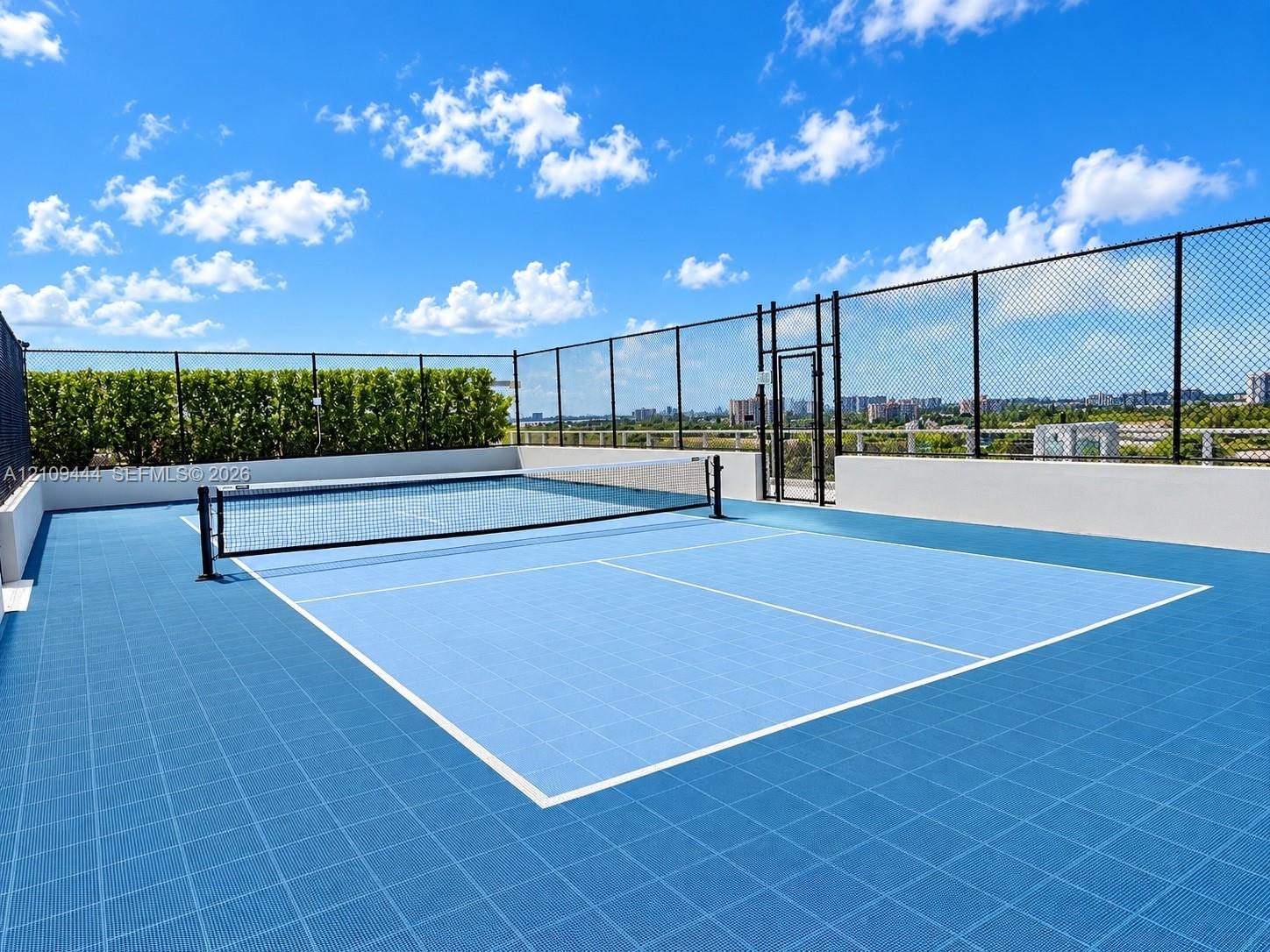 Rooftop athletic court with blue modular surface, perimeter chain-link fencing, and integrated privacy hedging