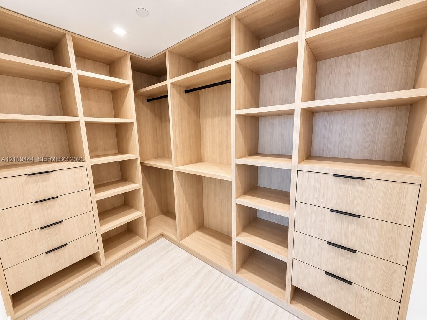 Custom walk-in storage featuring light wood-finish cabinetry, extensive built-in shelving, integrated garment rods, and two sets of drawers with matte